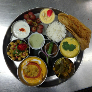 Our Awadhi Thali (clockwise from right): sultani dal, Awadhi-style okra dum, stuffed tomato in shahi gravy, sweet potato chaat, tomato-onion-cucumber raita, fruit, mango kulfi, basmati rice, parantha. Center: veg seekh kabab, hara bhara kabab, and special chutney.